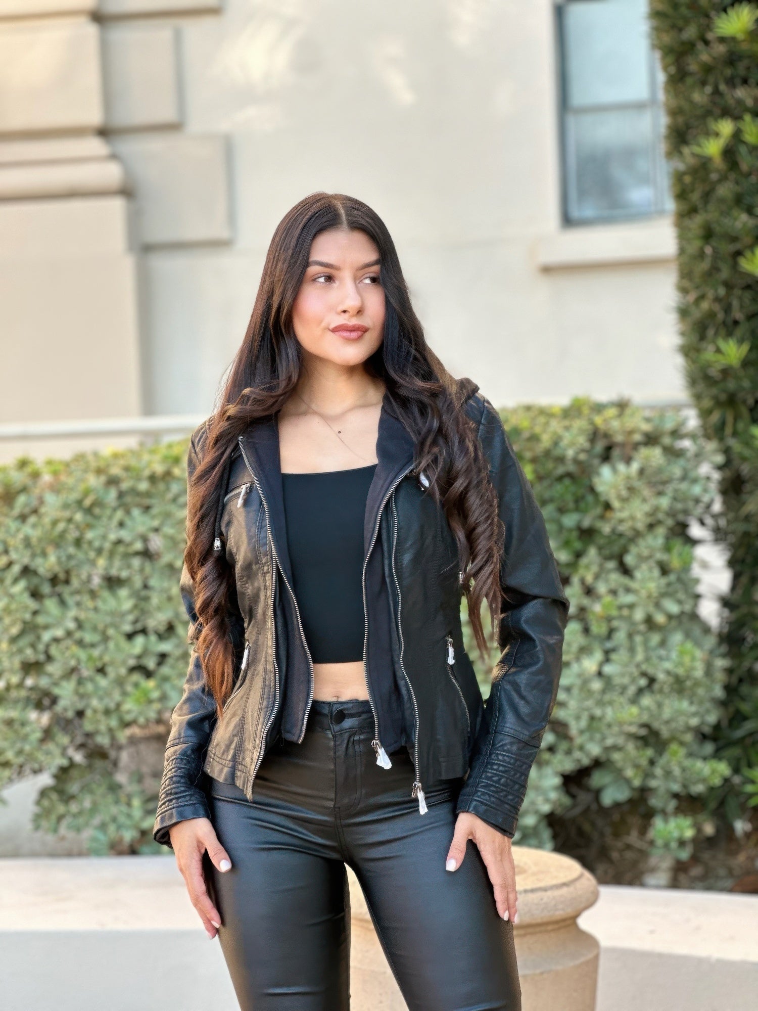 Women wearing a black leather jacket and pants standing outdoors with greenery in the background