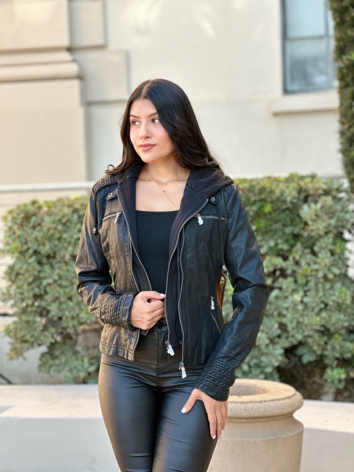 Women wearing a black leather jacket and pants standing outdoors with greenery in the background